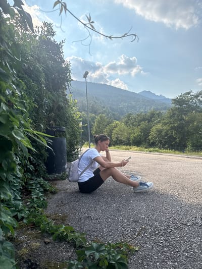 A woman is sitting on an asphalt road, holding a phone and looking at it. She is wearing a white t-shirt, black shorts, and blue and white sneakers. She has a light-colored bag slung across her chest. The woman has her legs bent and resting on the ground. In the background, there are trees and mountains. The sky is blue with some white clouds. The scene appears to be outdoors, possibly in a rural or mountainous area. The lighting suggests it is daytime, likely late afternoon or early evening, given the angle of the sun. There is no visible text in the image.