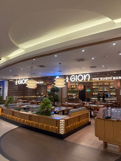 The image depicts the interior of a Gion The Sushi Bar restaurant located in Kelapa Dua, Indonesia. The restaurant's signage is prominently displayed above the dining area. The interior features a modern design with wooden furniture and decorative elements. Plants are incorporated into the decor. Patrons and staff can be seen within the restaurant.