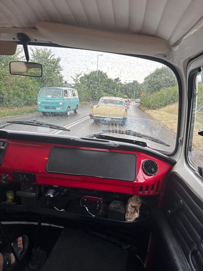 The image is taken from inside a vehicle, looking forward through the windshield. Raindrops are visible on the glass. The dashboard of the vehicle in the foreground is red with black accents and a black textured panel. A gear shift indicator with R, 1, 2, 3, and 4 is visible on the red dashboard. To the right of the red dashboard is a black vent. Below the dashboard is a black compartment with a brown paper bag and other items. A person's left foot wearing a black sandal is visible on the floor to the lower left.
Outside the vehicle, on a wet asphalt road, several cars are driving in a procession. The closest vehicle ahead is a yellow and blue vintage car. Further ahead is a turquoise vintage van. Trees and vegetation line the sides of the road. The sky appears overcast. The scene suggests a vintage car rally or parade.
The location is identified as Falkenberg, Sweden.