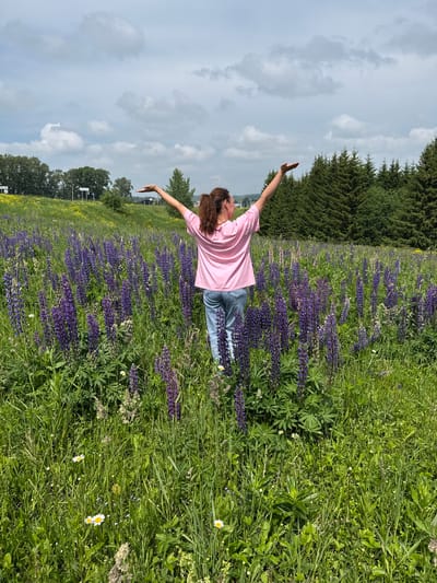 A young woman with brown hair pulled back in a ponytail stands in a field of purple lupine flowers, facing away from the camera. She wears a pink t-shirt and blue jeans. Her arms are raised to the sides with her palms facing upwards, as if embracing the environment. The field of lupines extends into the background, where it meets a line of evergreen trees. The sky is cloudy and a light blue. The location is the city of Kliuchiki, Russia.