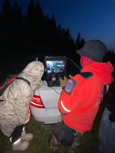 In the village of Perebor, Russia, at dusk, two individuals are gathered around the open trunk of a white car. On the trunk, a ruggedized tablet displays a drone's live feed, showing what appears to be a thermal image of a wooded area. One person, wearing a camouflage jacket and light-colored boots, leans over the tablet, while the other, in a bright red jacket and a black bucket hat, controls a device. The red jacket features a circular patch with Russian text and a graphic of a search and rescue dog. The surrounding environment is characterized by dark trees and a twilight sky.
