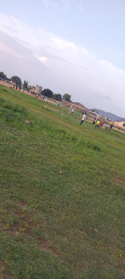 The image depicts a soccer game in progress on a grassy field, likely in a rural or semi-rural setting in or near Ibo, Nigeria. The field is uneven with some patches of bare earth visible in the foreground. A soccer goal is positioned in the mid-ground with players surrounding it, some wearing bright colored shirts. Other individuals are standing on the sidelines. In the background are buildings, trees, and a low mountain range. The sky is partly cloudy.