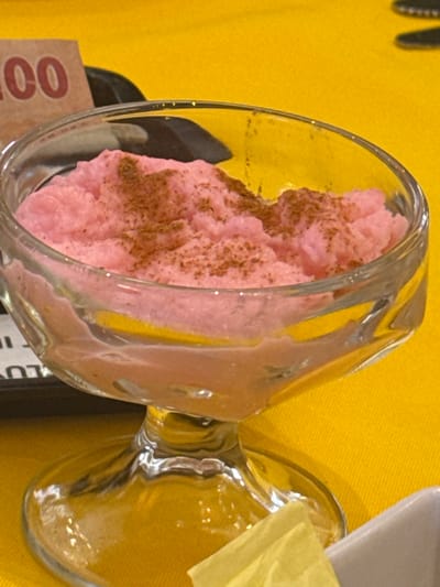 A glass of pink dessert, possibly ice cream or mousse, dusted with brown powder, is on a yellow surface, possibly a tablecloth, in an indoor setting. The glass is partially transparent, and its contents are visible. Cutlery is vaguely visible in the background on the left. Other objects and surfaces are also present but not fully identifiable due to the shallow depth of field and composition. The scene does not provide any information to connect it to the city of San Cristóbal, Mexico.