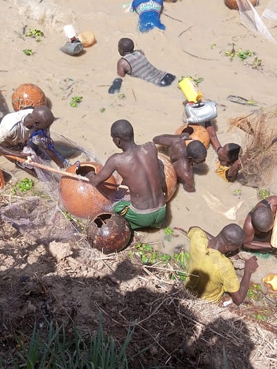 In Kokani North, Nigeria, a group of at least seven individuals is depicted engaged in activity within shallow, turbid, muddy water adjacent to a dirt bank. The scene shows multiple dark-skinned males, most partially submerged, appearing to be fishing or collecting.

In the foreground, a shirtless male wearing green shorts kneels in the water, facing away from the camera, interacting with a large, terracotta-colored pot or jar. To his left, another male in a white t-shirt manipulates a fishing net near a similar pot. A smaller, dark brown, spherical object with a hole is visible in the water near the green-shorted individual.

Further into the water, another male wearing a striped sleeveless top is submerged from the waist down, appearing to be moving through the mud. Behind him, a blue garment, possibly a shirt, is partially submerged. Several other males are distributed across the scene, some shirtless, others wearing yellow or light-colored shirts, all in various states of immersion and interaction with the water, nets, and containers.

Floating objects in the muddy water include various plastic containers (white, yellow, silver) and multiple large, spherical, netted objects, possibly gourds or floats. Sparse green aquatic vegetation is scattered throughout the water. The immediate foreground consists of a dry dirt bank with some patchy green grass and distinct shadows, indicating bright sunlight.