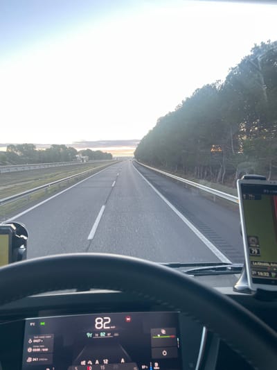 A driver's perspective from inside a vehicle speeding down a straight highway. The speedometer reads 82 km/h. A navigation device shows a route towards "La Almolda". The car's dashboard indicates a 5°C outside temperature and 37% battery level. Roadside trees and a clear sky are visible.