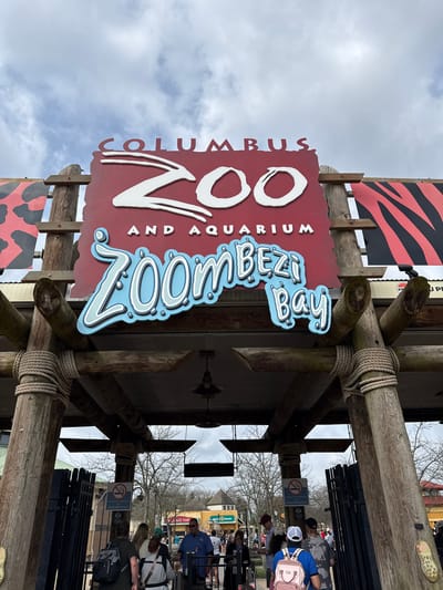 Here's a description of the image:
The image shows the entrance sign to the Columbus Zoo and Aquarium in Powell, Ohio, United States of America. The sign is made of a red background with the words "Columbus Zoo and Aquarium" written in white letters. The words "Zoombezi Bay" are written in blue letters with white accents on a separate sign attached below. The sign is attached to a wooden structure made of logs and rope. In the background, there are people lined up to enter the zoo. There is a sign that reads "Smoke-Free Facility." The sky is overcast.