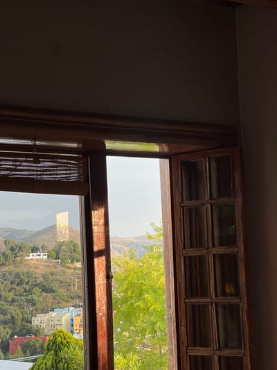 A view from inside a room looks out through a dark wood-framed window onto a hillside cityscape in Guanajuato City, Mexico. The upper section of the image shows the dark interior ceiling and wall. The window frame, consisting of a lintel, sill, and vertical mullions, is dark wood, with direct sunlight illuminating its left vertical and horizontal elements. A horizontal slat-style blind is partially lowered over the left pane of the window. The right side of the window is open, with a multi-paned wooden shutter or door swung inwards to the right.

Beyond the window, a bright, hazy sky is visible above a landscape of rolling hills covered in dense green foliage. In the mid-ground, a cluster of brightly colored buildings—yellow, blue, and red—are visible below the tree line. Further back, on a more distant hill, a distinct, tall, light-colored rectangular monument, identified as the Monumento al Pípila, stands prominently. A white house is situated on a closer, vegetated hillside to the left of the monument. No individuals, specific actions, or interactions are present in the scene.