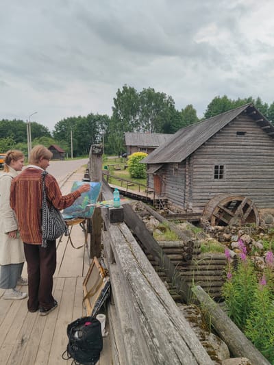Two individuals, one with blonde hair in a light-colored coat and grey pants, and another with blonde hair in a striped orange and brown shirt and dark pants, stand on a wooden walkway. The person in the striped shirt is painting on an easel, holding a paintbrush and palette. A water bottle and other art supplies are visible on the walkway. Behind them, a rustic wooden building with a water wheel is situated next to a rocky, overgrown area with pink flowers. Trees and a road are visible in the background. The overall setting appears to be an outdoor, rural environment, possibly an historical village like Bugrovo village in Russia. The sky is overcast.