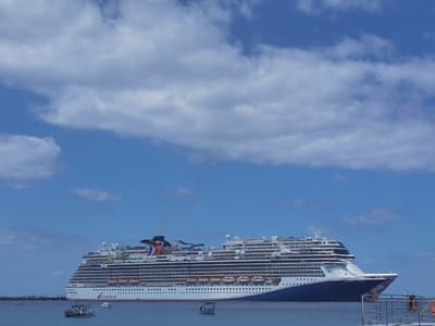 The image shows a large cruise ship docked at the pier in Cozumel, Mexico. The ship is white with blue and red accents. The name "Carnival" is visible on the side of the ship. The ship is docked in the water, and there is a small boat in the foreground. The sky is blue with white clouds. The coordinates (latitude: 20.512, longitude: -86.95) place the location in the port area of Cozumel. The scene is peaceful and serene.
