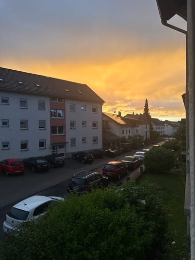 The image captures a residential street scene in Idstein, Germany, viewed from an elevated perspective, likely a balcony or window, during sunset. The sky, dominating the upper right portion of the frame, transitions from a vivid golden-orange hue to a pale blue-grey on the left, indicating the atmospheric conditions of either sunrise or, more likely, sunset.
In the midground, a row of multi-story residential buildings lines the street. The closest building features a white facade with numerous square windows, some open, and a distinctive reddish-orange painted section on one floor. Its roof is covered with dark tiles. Further down the street, more buildings with similar architecture are visible, along with a prominent, tall, slender coniferous tree rising above them.
The street itself is paved and lined with numerous parked cars of various models and colors, including red, black, white, and silver. Lush green bushes and trees flank the street, and a grassy area is visible to the right, adding natural elements to the urban setting.
In the immediate foreground, a dark, metallic railing or balcony structure extends across the bottom of the frame, partially obscuring the view. A dark gutter and a section of a building's roof are also visible in the bottom-left corner.
The scene is quiet, with no people visible or specific activities taking place, conveying a sense of tranquility. The warm, soft light from the setting sun casts long shadows and imbues the entire landscape with a peaceful and serene atmosphere. Visible text includes "ID 100..." on the rear license plate of a white car and "ID 30..." on a red car, with "ID" being the vehicle registration code for the Idstein district.