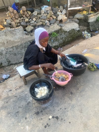 The image depicts an outdoor scene in Idon Kasa, Nigeria, focusing on a woman engaged in food preparation. The foreground features a woman of African descent, wearing a black long-sleeved garment, a white headscarf over a maroon underscarf, a gold-colored necklace, and multiple bracelets on both wrists. She is seated on a small, light-colored wooden stool. In front of her, a black cooking pot or basin containing oil and several square, white pieces (likely tofu or yam) is actively frying, producing visible bubbles. This pot rests on a low, dark-colored stand, suggesting a charcoal stove. To her right, a pink plastic colander holds numerous golden-brown, fried pieces of food, matching those in the cooking pot. She holds a metal fork or spatula, gesturing towards the colander. Adjacent to the colander is a large, black plastic basin containing various items, including multiple black plastic bags, a white plate, and other indistinct materials.
The ground is concrete and shows dark stains, possibly from oil or grease. In the mid-ground, a concrete barrier or drainage channel runs horizontally. Behind this barrier, a substantial pile of rough, irregular rocks and stones is stacked. To the left of this pile, partially obscured, another individual is visible, wearing a blue garment and a light-colored cap, seated. Further into the background, beyond the rocks, corrugated metal sheeting and additional debris are present. The lighting suggests daytime, with shadows indicating a low sun angle.