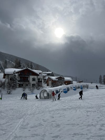 The image depicts a snow-covered ski area in Davos, Switzerland, under an overcast sky with a prominent, bright sun partially visible through the clouds. Light snowfall is observable. In the midground, a group of individuals engages in winter sports on a gentle slope.

From left to right, on the snow, two individuals on skis, dressed in winter gear and helmets, stand facing each other. To their right, near a covered conveyor belt, a child wearing a light green helmet is seated on the snow. An adult in a red jacket and light pants, on skis, stands near the entrance of a transparent, tunnel-like covered conveyor belt lift, appearing to assist a child on skis who is partially inside the entrance. The conveyor belt structure is heavily covered in snow and features blue elements visible through openings on its side. Further to the right, a person in dark clothing and a backpack walks uphill on the snow, using ski poles. In the distant background on the right, two additional figures are visible walking on the snow.

The background features multiple multi-story buildings, typical alpine chalets with dark wood facades and white plaster, all with snow-laden pitched roofs. Pine trees, also covered in snow, are interspersed among the buildings and ascend the mountain slope on the left. A ski lift tower is partially visible on the far left, and a ski lift line with chairs is seen in the distance on the right, extending towards a snow-covered ridge. Ski tracks are visible on the foreground snow.