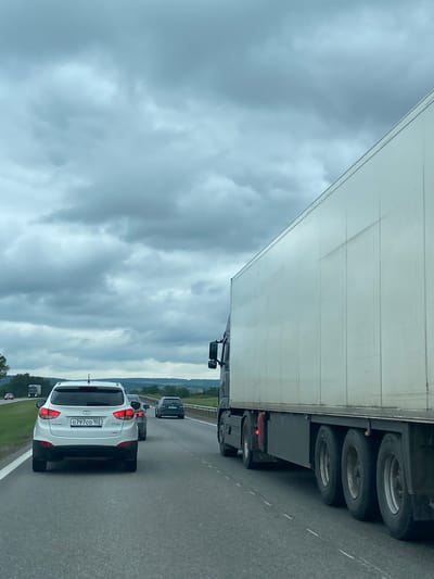 The image depicts a cloudy day on a highway in Uzytamak, Russia. The foreground shows the rear of a white Hyundai SUV with a license plate reading "0797co 102". To the right of the SUV, a large white semi-trailer truck occupies a significant portion of the frame, its trailer extending towards the right edge. In the middle ground, other vehicles, including a dark-colored sedan, are visible on the highway. The background consists of a tree line and rolling hills under a heavily overcast sky. The highway appears to have multiple lanes, indicated by dashed white lines.