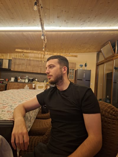 A man is seated in a chair, turned to his right, looking away from the camera. He has short, dark hair and a beard. He is wearing a dark, ribbed t-shirt. His left arm rests on a table covered with a patterned tablecloth. In the background, a kitchen is visible, featuring wooden cabinets, a refrigerator, and a stove. The room has a wooden ceiling with exposed light fixtures. The scene appears to be set in a residential interior, possibly a kitchen or dining area, in the city of Каменар, Bulgaria.