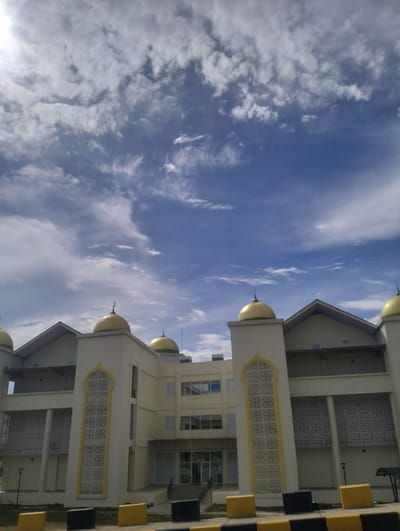 The image depicts a modern, cream-colored building with golden domes, likely a mosque, situated under a bright, partly cloudy sky. The building has multiple stories, with visible windows and ornate white latticework panels framed in yellow. The foreground shows a few safety barriers. The scene appears to be located in Reuleut Timu, Indonesia, as indicated by the architectural style and environmental context which is typical for the region. There are no people visible in the frame. The overall impression is one of a contemporary religious structure in an urban or semi-urban setting.