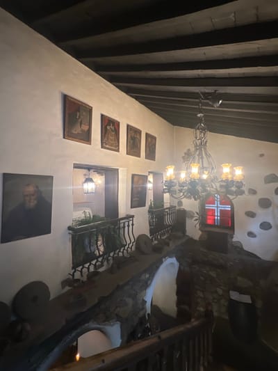 This media file depicts an interior scene, likely a rustic building, possibly a restaurant or historic home, given the stone walls, exposed wooden beams on the ceiling, and decorative chandelier. The lighting is warm and atmospheric, suggesting it could be evening or an interior space with artificial light.

The primary subjects are the architectural elements and decorations. A large, ornate chandelier with multiple lights hangs from the ceiling, casting a warm glow. Several framed artworks, appearing to be portraits or historical depictions, are hung on a textured white wall. A decorative iron railing with a black metal railing runs along an upper level or balcony, with greenery visible on the other side. A staircase with wooden steps and railings is also visible.

In the background, a window with red panes and a white cross design is visible, adding a unique visual element. The overall aesthetic suggests a traditional or historical setting, possibly with a touch of gothic or medieval influence due to the stonework and chandelier. The scene is static, with no apparent activity or event taking place.

Notable details include the rustic textures of the stone and wood, the vintage style of the chandelier and artwork, and the distinctive window design. The lighting creates a cozy and somewhat dramatic ambiance. The location is identified as La Vera, Spain, which could imply a regional architectural style or historical context. No visible text is present in the media.
