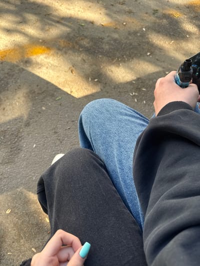 An overhead shot depicts a person seated outdoors, possibly on the ground in Severnaya Samarka, Russia. The individual wears a dark-colored hooded sweatshirt and blue denim jeans on one leg, while the other leg is clad in dark-colored pants. The person holds a vaping device in their left hand. The ground is a light-colored surface. A glimpse of a hand with a light blue nail is visible near the person's right leg.