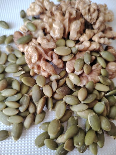 The image displays a pile of walnuts and pumpkin seeds on a white, textured surface. The walnuts are broken into several pieces, revealing their characteristic wrinkled texture and light brown color. Scattered amongst and beneath the walnuts are numerous green pumpkin seeds, which are oval-shaped and have a smooth surface. Some of the pumpkin seeds are slightly lighter green, and a few have a darker, more brownish hue. The lighting appears to be natural and diffuse, casting soft shadows. The scene is a close-up shot, focusing entirely on the food items. There are no people or animals visible. The overall impression is of a healthy, natural snack or ingredient. The location context provided is Кривец, Russia, but there are no specific visual cues in the image to confirm this location.