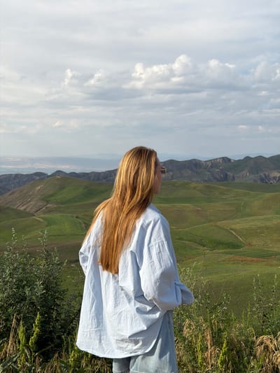A woman is captured in a medium shot, looking out at a vast landscape. She is seen from behind and to the side, with her long, blonde hair flowing down her back. She is wearing a light blue button-down shirt and blue jeans. Her posture suggests contemplation or relaxation as she takes in the view.

The setting is a lush, green mountainous region, likely in Toguz-Bulak, Kyrgyzstan. Rolling hills dominate the background, with visible textures and contours suggesting a natural, untamed environment. The scale of the mountains and the open sky create a sense of grandeur and isolation.

The sky above is a mix of white and gray clouds, indicating a partly cloudy day. The natural light suggests it could be late morning or early afternoon. The overall mood is serene and peaceful, with the woman appearing to be enjoying a moment of quiet reflection amidst the natural beauty. The foreground features dry grass and some green foliage, adding to the natural aesthetic. There is no visible text in the image.