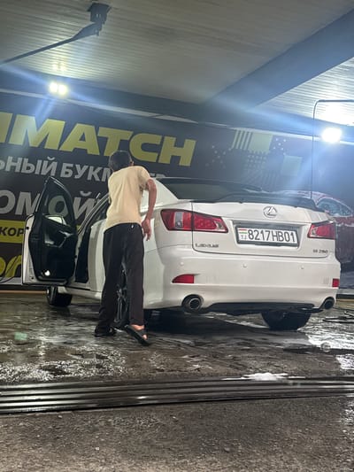 A young man wearing a light-colored t-shirt and dark pants is standing at the open driver's side door of a white Lexus IS 350. The car's license plate reads "TJ 8217 HB01". The scene appears to be an outdoor car wash with a wet, foamy concrete floor. In the background, a large banner with yellow lettering that says "MMATCH" is visible. Another red car is partially visible in the background, as well as some lighting fixtures. The image was taken in Пистамазор, Tajikistan.