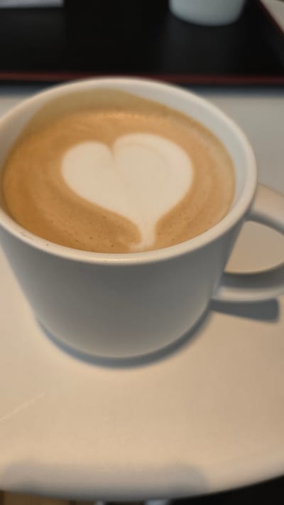 A close-up, eye-level shot shows a white ceramic mug filled with latte art, a heart shape formed by milk foam on top of the coffee. The mug rests on a white saucer. In the background, out of focus, is a dark surface and a sliver of another white mug. The scene is likely indoors, possibly in a cafe in Gwangmyeong, South Korea.