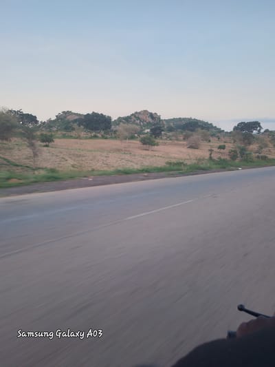 The image depicts a rural landscape, possibly near Jibilik, Nigeria, with a paved road stretching into the distance. The road shows motion blur, suggesting it was taken from a moving vehicle. On the left side of the road, a field with rows of planted crops is visible, bordered by sparse vegetation and trees. In the background, gently rolling hills are covered with trees and rocky outcrops. The sky is clear with a pale blue hue, indicating either early morning or late afternoon light. The text "Samsung Galaxy A03" is superimposed on the lower left of the image, identifying the device used to capture the photograph. A portion of a hand and handlebars, likely from a motorcycle or bicycle, are visible in the lower right corner, further confirming the perspective of a moving vehicle.