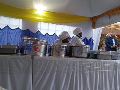 The image captures a food service setup under a tent at an event in Shimbi Mashariki, Tanzania. In the foreground, a buffet table draped with a white tablecloth holds several stainless steel food warmers and chafing dishes, along with a stack of white plates. One prominent food warmer clearly displays the brand name "Maxima."
Two women, dressed in white chef hats and matching white uniforms, are positioned behind the table, seemingly preparing for or attending to the food service. Their gaze is directed generally forward or slightly to the right, indicating their role in the event.
In the background, to the right, a man in a dark blue suit is visible, looking down at an item in his hands, possibly a mobile phone. The setting is elaborately decorated, featuring a white tent structure with vibrant yellow fabric draped along the top and ruffled blue fabric along the lower walls. Bright light from above illuminates the scene, suggesting it is daytime or a very well-lit indoor tent space. The overall impression is a catered social gathering or celebration.