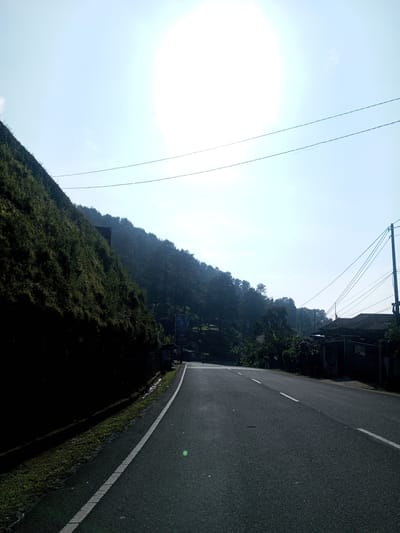 The image shows a two-lane asphalt road curving through a hilly landscape. On the left side of the road is a steep embankment covered in dense vegetation. A line of trees and more vegetation is visible further along the road, leading into the distance. A white line marks the edge of the road. On the right side, there is a building with a dark roof and foliage near the road. Power lines run across the sky. The sun is high and bright, creating a hazy appearance in the sky. The image lacks specific details to confirm its location in Phouviang-Gnai, Laos.