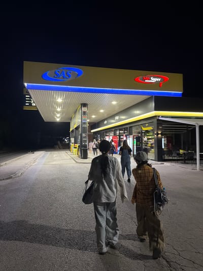 The image depicts a night scene at a SAS Super gas station in Mali Zvornik Municipality, Serbia. Two women are walking away from the camera on the asphalt surface of the gas station. They are walking towards a brightly lit building that appears to be a convenience store or shop, attached to the gas station. The building has a yellow and black facade with illuminated signage that reads "cafe" and "shop." Inside the shop, shelves stocked with various items are visible through the glass windows. The gas station canopy is illuminated with bright lights, and the SAS logo is prominently displayed in blue and red. Several cars are parked at the gas pumps. The overall environment is well-lit, suggesting the gas station is open for business at night.