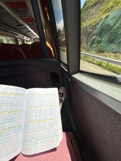 The image depicts the interior of a bus, with a person's legs and feet wearing black Converse sneakers visible in the lower right corner. An open notebook with handwritten notes in Bulgarian is placed on the person's lap. The notes appear to be related to legal or business terminology, with highlighted sections. Through the bus window on the right, a green, terraced hillside covered in vegetation is visible, suggesting a rural or mountainous setting. A guardrail runs along the edge of the road outside the bus. The lighting indicates it is daytime, with sunlight streaming into the bus. The location of Staro Selo, Bulgaria is not explicitly visible or derivable from the provided image.