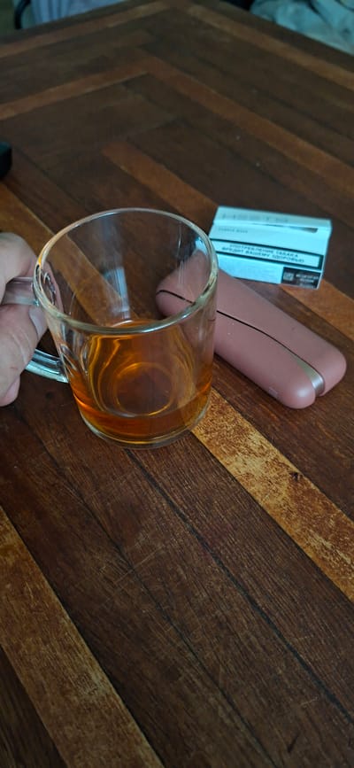 A close-up, eye-level shot shows a transparent glass mug filled with amber-colored liquid, resting on a dark wooden table with a herringbone pattern. A hand is visible on the left side of the mug, holding it. To the right of the mug, a pink electronic smoking device and a white pack of cigarettes are placed on the table. The background is blurred, showing more of the wooden table. The scene appears to be indoors. The image does not contain information about Shakhtarsk, Ukraine.