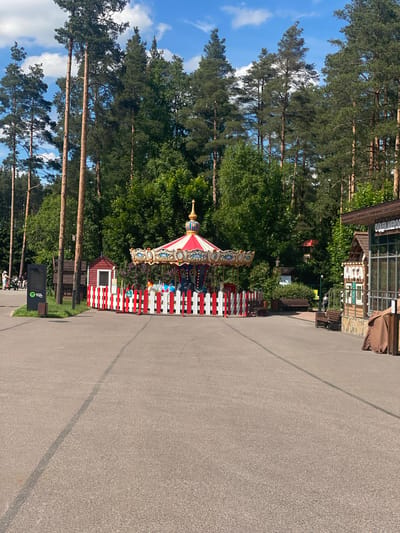 In Mistolovo, Russia, on a bright, sunny day, a carousel with red and white striped fencing stands in an open paved area, surrounded by tall pine trees and lush green foliage. A small red building with white trim is situated to the left of the carousel. In the foreground, a paved path stretches into the distance, marked by a dark line. To the right of the carousel, a wooden building with a sign that reads "ОТАЛМА МЕРОД" is visible, with a brown tarp draped over a portion of its exterior. Benches are placed strategically throughout the scene. A few individuals are seen in the distance on the left, near a digital information board. The sky above is a vibrant blue with scattered white clouds.