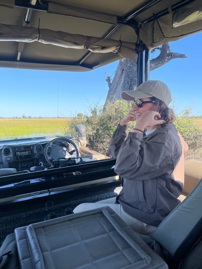 The image, taken at a sideways perspective, shows a person inside a safari vehicle in Kakoro, Botswana. The person, wearing a grey cap, sunglasses, and a grey jacket, is holding their glasses near their face. The vehicle's interior includes a steering wheel, dashboard with various controls, and grey seats. Through the open side of the vehicle, a grassy plain stretches under a clear blue sky, with a tree visible in the background. The frame of the safari vehicle, including the canvas roof, is visible on the left side of the image.