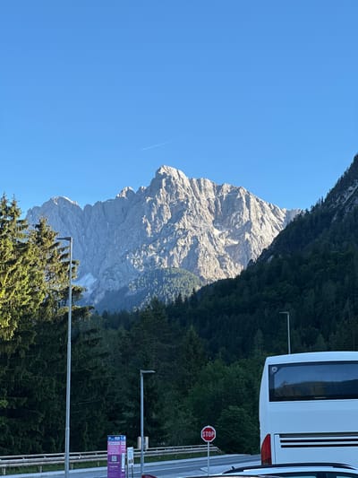 The image shows a scenic view of the Julian Alps in Kranjska Gora, Slovenia, on a clear, sunny day. In the foreground, a white tour bus is parked, its rear visible on the right side of the frame. Adjacent to the bus, a stop sign is posted. Further to the left, a row of trees, including tall coniferous trees, lines the foreground, with a tall lamppost also visible. In the mid-ground, a dense forest of dark green trees covers the lower slopes of the mountains. The main focus of the image is the towering, jagged mountain range of the Julian Alps, bathed in sunlight, revealing its rocky, textured surface. A thin contrail from an airplane stretches across the clear blue sky above the mountains.