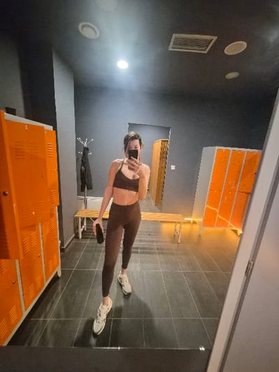 A female individual is captured in a mirror selfie within a locker room or changing facility, presumed to be located in the Municipal Unit of Corinth, Greece. She wears a dark brown sports bra, matching dark brown leggings, and white and beige athletic shoes. Her face is obscured by a black smartphone she holds to take the photograph. The room features dark gray walls and a dark gray tiled floor. Numerous orange metal lockers are visible on both the left and right sides of the space. A light-colored wooden bench runs across the background, behind which a black coat rack holds several dark garments. The ceiling is dark gray with recessed circular lights and a rectangular ventilation grate.