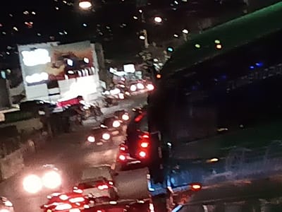 A night scene captures a busy street with traffic. Car headlights and taillights illuminate the road, indicating movement. A large building with illuminated signage dominates the left side. An overpass structure is visible to the right, with lights indicating its presence. The background features scattered lights, potentially from other buildings or streetlights. The image is slightly blurry, suggesting it may have been taken from a distance or with a moving camera.