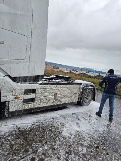 A male individual is observed from behind, actively pressure-washing a large white truck outdoors in an industrial or commercial service area in Ostend, Belgium. The truck, a tractor unit, is positioned on the left and center, largely covered in white soap foam, with underlying dirt visible on its lower chassis, wheels, and side steps. The individual wears a dark beanie, a dark blue jacket (with partially visible white text on the back), blue jeans, and dark shoes, holding a pressure washer lance aimed at the truck's rear wheel and lower side. The ground is a dark, wet surface extensively covered in white soap foam, with visible footprints. A dark hose from the pressure washer lies on the ground in the foreground. The background features a grassy area, a fence, and distant industrial-style buildings with light-colored roofs, all beneath an overcast, gray sky.