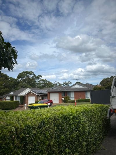 A residential street scene under a cloudy sky shows row houses with brick facades, some featuring garages and solar panels. A red and yellow car is parked in front of one of the houses. Lush green hedges line the street, partially obscuring the houses. A utility vehicle is partially visible at the top of the frame. Some trees are interspersed between the houses, and foliage is visible in the foreground.