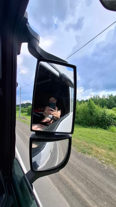 The image is a reflection in a truck's side mirror. Inside the truck cab, a person wearing glasses and a gray tank top is taking a selfie with a smartphone. The person has short hair and is giving a peace sign. They are sitting in the driver's seat. Outside the truck, a rural landscape with green trees and grass is visible, along with a dirt road. The sky is partly cloudy. The image was captured in Sulenka, Russia.