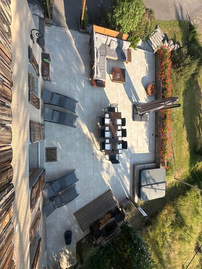 An elevated, downward-angled view depicts an unoccupied outdoor patio area situated beside a light-colored building in Saint-Jean-d'Aulps, France. The patio floor consists of light-colored, square paving tiles, illuminated by direct sunlight casting distinct, long shadows.
In the upper right, an L-shaped outdoor lounge sofa with light cushions and two wooden coffee tables are present; one small table holds unidentified items including a green object and dark rectangular objects. A dark rectangular dining table, set for twelve with white plates and glassware, occupies the central area, surrounded by chairs. Adjacent to the dining table on the right, a large, closed brown patio umbrella is positioned. A red flower bed borders the patio's right edge, extending alongside a green lawn. A large, round wooden block is situated near the flower bed.
In the bottom right, a covered hot tub rests on a wooden deck. Towards the bottom center, an outdoor barbecue station with firewood storage is visible on another wooden platform. A dark cylindrical waste bin is positioned near the barbecue. Two pairs of dark sun loungers are arranged on the left side of the patio, closer to the building wall. A square metal utility cover is visible in the patio floor. The adjacent building wall features multiple windows with open and closed wooden shutters, and a black wall-mounted lantern is mounted near an upper window. No individuals are present within the scene.