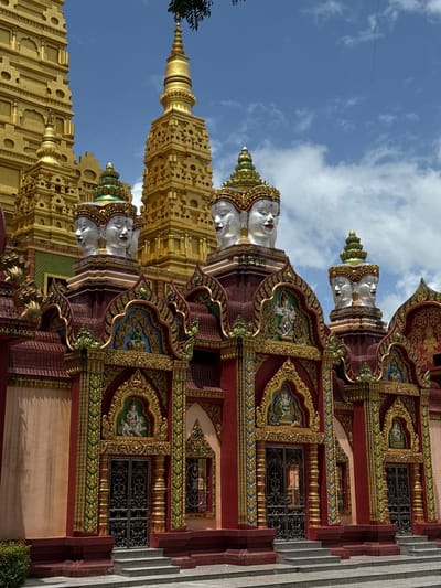 The image depicts the exterior of a temple in Na Nuea, Thailand. The temple features a striking facade with ornate architecture and religious iconography. Prominently displayed are several statues of multi-faced heads, with some appearing to have four faces each. These heads are white with red lips and are adorned with golden headdresses.
Behind the facade, tall, golden stupas rise into a bright blue sky dotted with white clouds. The lower section of the temple is painted in rich colors, predominantly red and a muted pinkish-orange, with intricate gold and green detailing. Arched doorways with decorative metal gates are visible, and above each doorway, there are recessed niches containing figures. Steps lead up to the entrances. The overall impression is one of grandeur and spiritual significance.