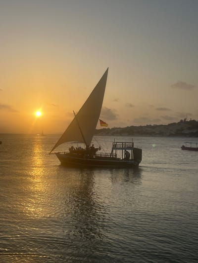 A sailboat filled with people glides through golden water under a setting sun. The light reflects brightly off the water's surface, creating a dazzling path. A small flag flutters atop the boat's mast. A nearby landmass is visible in the background, and another small boat is seen further away on the water. The sky is a blend of pale blues and yellows.