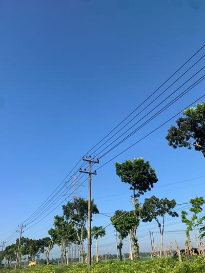 The image depicts a rural scene in Karangkemiri, Indonesia, under a clear blue sky. Multiple utility poles with power lines traverse the frame, stretching from the left foreground towards the upper right corner. A line of trees, varying in height and foliage, runs horizontally across the mid-ground, interspersed with more utility poles and distant, larger transmission towers. The foreground is dominated by a field of lush, green crops, possibly agriculture. In the far left of the frame, a yellow piece of agricultural machinery, likely a tractor, is visible. The overall impression is one of a working agricultural landscape integrated with essential infrastructure.