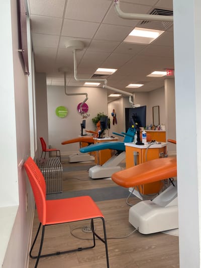 This is an indoor photograph of a dental office in Chesapeake, Virginia, United States of America. The office appears to be modern and brightly lit, with multiple dental chairs. In the foreground, there is a bright orange plastic chair with black metal legs, leaning against a white wall. To the right of this chair, a metal bench is visible.
Further into the room, there are several dental chairs. Two are prominently visible: one is teal blue, and another is orange. These chairs are situated at dental workstations equipped with monitors and cabinetry. The workstations are predominantly orange. In the background, there are additional dental chairs, though less distinct.
The ceiling is a suspended tile grid with multiple fluorescent light panels. Several articulated medical lights hang from the ceiling, positioned above the dental chairs. On the wall, there are circular decorative elements, one green and one purple, with text that is partially obscured. An "EXIT" sign with red lettering is visible on the far right of the image, mounted near the ceiling. The flooring appears to be wood-look laminate. There are no people visible in the photograph.