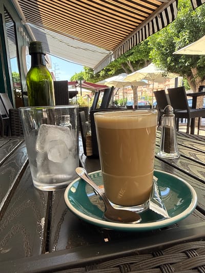 The image depicts a close-up, horizontally oriented view of a table at an outdoor cafe. On the table, which appears to be made of dark wood with a textured surface, are several items. In the foreground to the left is a green glass bottle lying on its side, partially obscured by a glass of ice water. The glass of ice water is in focus, with large ice cubes visible within. To the right of the ice water is a glass with a light brown, frothy beverage, likely coffee or a similar drink. This glass sits on a dark green saucer, which also holds a small packet of sugar and a spoon. Behind the drinks, a salt and pepper shaker with a metallic top is visible.

In the background, the scene suggests a sunny day at an outdoor dining area. Striped awnings in black and tan, along with white umbrellas, provide shade. Green foliage from trees is also visible, suggesting a lush outdoor environment. The overall setting appears to be a relaxed cafe or restaurant, possibly in Campillo Del Moro, Spain, given the presence of outdoor seating and umbrellas typical of Mediterranean climates. The lighting suggests it is daytime, with the sun casting shadows and highlighting the textures of the table and drinks. No people are clearly visible, but the presence of table settings implies patrons were recently present or are expected. The image focuses on the beverages and the table setting, creating an inviting, serene atmosphere.