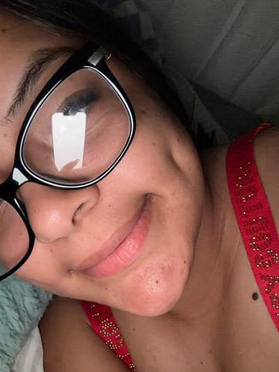 This is a close-up, portrait-oriented, selfie-style photograph of a person's face. The subject is wearing black-rimmed glasses and a red top with gold embellishments. The subject's left eye is visible through the glasses, with a reflection of a smartphone screen visible on the lens. The subject has dimples and a mole on their chin. The background is dark and appears to be an indoor setting, possibly a bed with a dark blanket or comforter. There is no indication of the city of Balch Springs, United States of America, in this image.