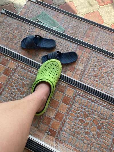 A first-person perspective shot shows a person's bare leg and foot wearing a bright green Croc shoe. The shoe is positioned on an outdoor step with a textured tile surface. In the background, on the same step, are two dark blue flip-flops. Above these, on another step, is a folded light green towel. The steps appear to be part of an exterior pathway, likely leading to a building. The environment suggests a warm, sunny day. The image does not provide enough detail to determine a specific location within the city of Kursk, Russia.
