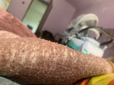 The image shows a close-up view of a brown knitted fabric, possibly a blanket or sweater, with visible individual fibers and a subtle pattern of lighter colored threads woven in. In the background, out of focus, a room is visible with purple walls. A doorway with a light-colored door is seen on the left, and on the right, a baby stroller or carrier with a white canopy is present. The scene appears to be indoors, in a domestic setting. No specific location within Krushev Dol, Bulgaria, can be determined from the image.