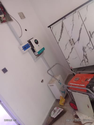 The image depicts an indoor scene, likely a utility room or a space under construction. The walls are painted white. On the left wall, an electrical panel is mounted, with wires leading from it. Below the panel, a white device with blue buttons is attached to the wall. To the right of this, a larger white unit with a circular display showing "32.0" is mounted, possibly an inverter or a control unit.
In the foreground and extending into the middle ground, various objects are present on the floor. These include a white rectangular appliance, a bright yellow bag, and a red wheeled object which appears to be part of a generator or compressor. A piece of paper or packaging with text is also visible on the floor. The background features a wall with large marble-patterned tiles, suggesting a finished or partially finished area.
The lighting suggests it is daytime, but the specific time is indicated by the text overlay at the bottom right: "06/17/2026 15:01". The text "REDMI A3" is also visible in the bottom left corner, indicating the device used to capture the image. No people are visible in the frame. The scene appears to be utilitarian and possibly related to electrical installations or equipment setup, with a mix of unfinished and finished elements. The location context is Nwofia, Nigeria.