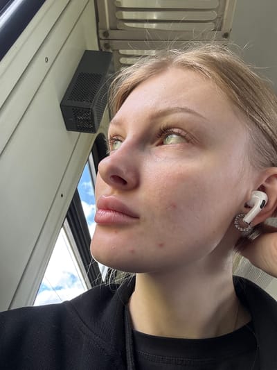 A close-up, slightly angled photograph depicts a young woman with fair skin, light blonde hair, and light-colored eyes, possibly green. She is wearing a white earbud in her left ear, adorned with a circular, possibly jeweled, earring attachment. She is wearing a black shirt, and a delicate silver chain is visible around her neck.
The environment appears to be an interior space with white or light-colored metal panels. A small, rectangular black object with a perforated surface rests on the panel to her left. Through a window, a glimpse of a blue sky with white clouds is visible. The image doesn't provide any additional information that would definitively place this scene in Mysy, Russia.